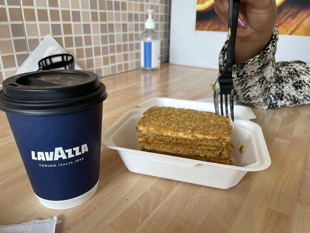 A takeaway coffee cup beside a slice of honey cake in a box on a wooden cafe table