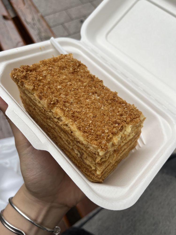 A hand holding an open takeaway box with a thick slice of layered honey cake topped with crumb